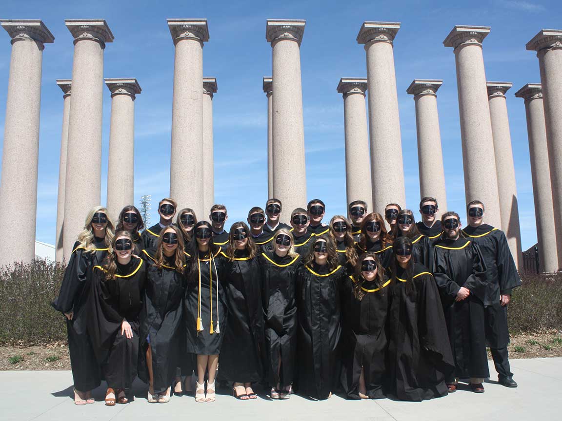 students in black gowns in front of pillars