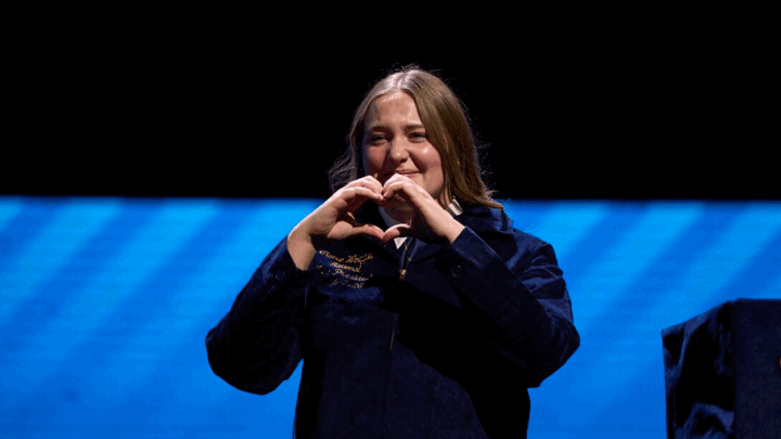 Young woman in an FFA jacket standing on stage, smiling and forming a heart shape with her hands against a blue-lit background.